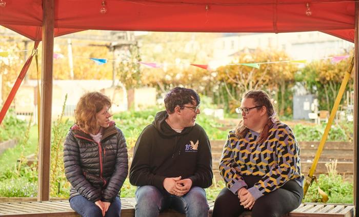 three young people are chatting together, outside in the museum's garden. They're sitting on a wooden bench under an orange canopy.